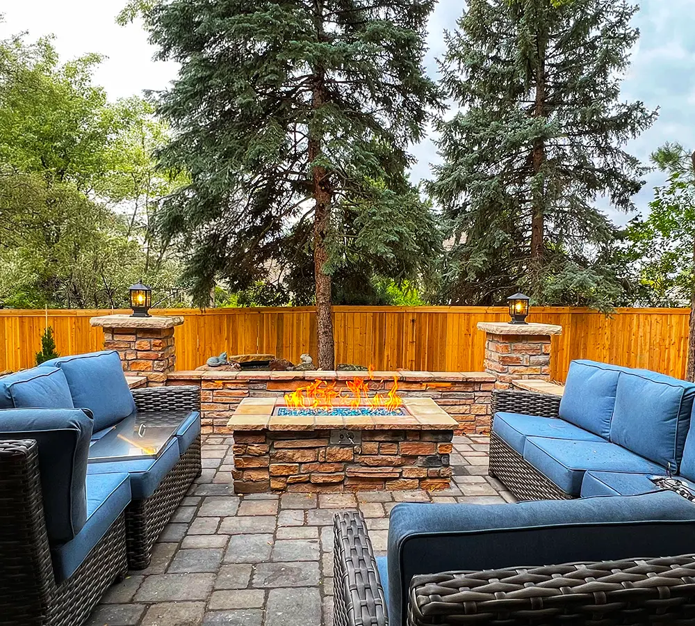 Cozy outdoor patio with stone fire pit, blue cushioned wicker seating, and surrounding trees for a relaxing backyard retreat.