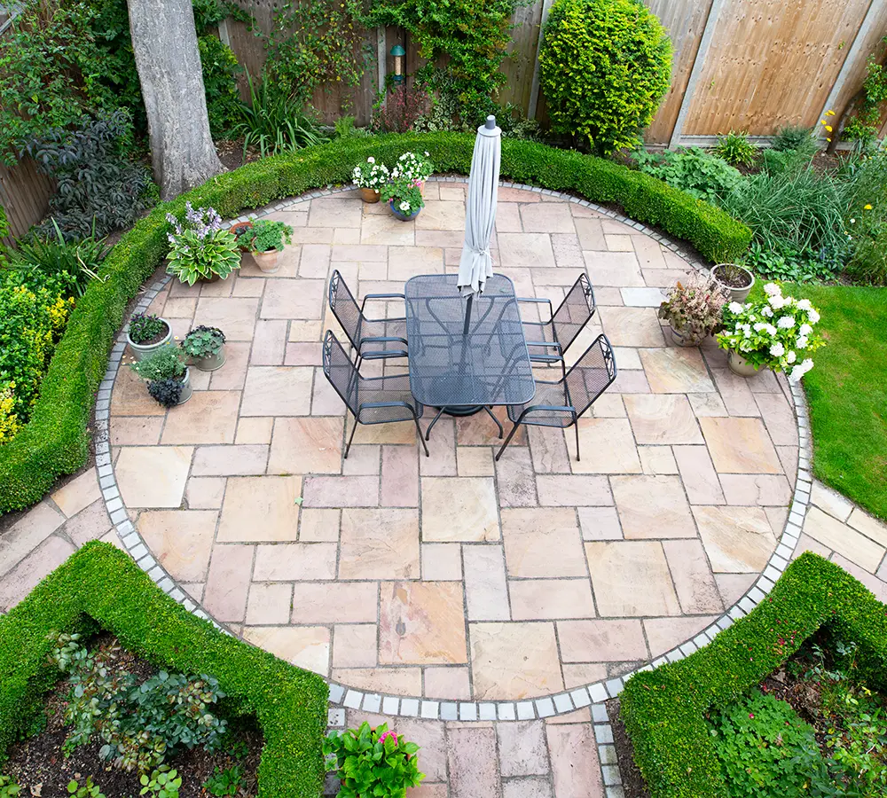 Circular patio with elegant stone pavers and dining set, surrounded by lush greenery in a backyard garden setting.