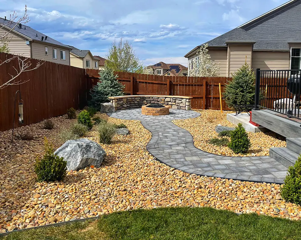 Backyard landscaping with stone path leading to fire pit, surrounded by decorative rocks and greenery.