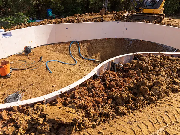 In-ground swimming pool installation site with excavation, plumbing installation, and heavy equipment in progress.