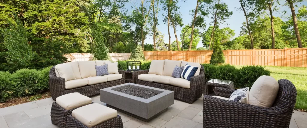 A stylish backyard patio with a fire pit, wicker seating with cream cushions, and lush greenery enclosed by a wooden fence
