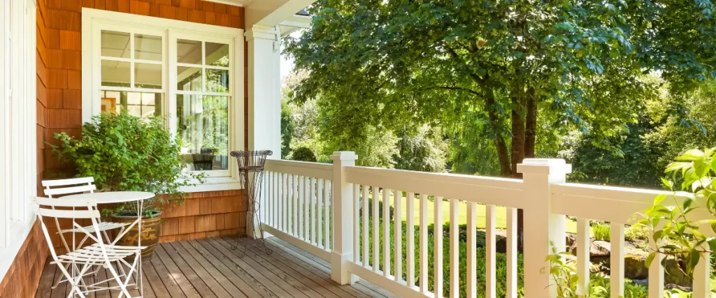 A charming porch with white fencing, wood flooring, a small bistro set, and lush green trees providing shade