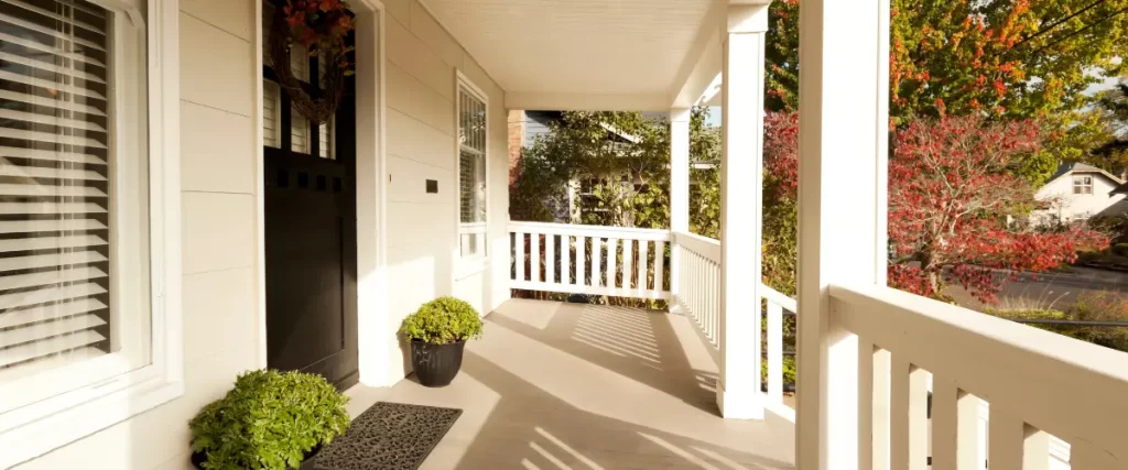 A welcoming front porch with white railings, a black door, potted plants, and autumn foliage in the background