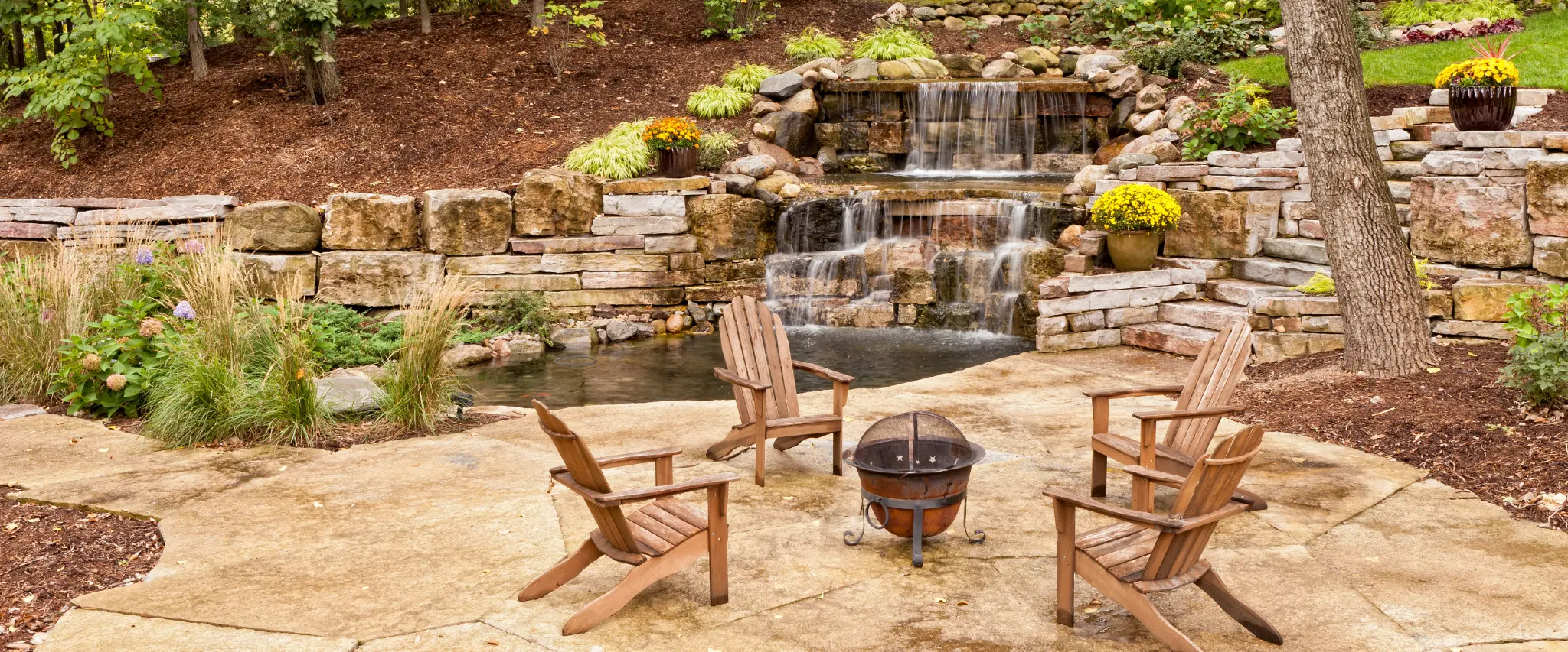 A backyard patio with Adirondack chairs, a built-in firepit, and a cascading waterfall surrounded by natural stones.